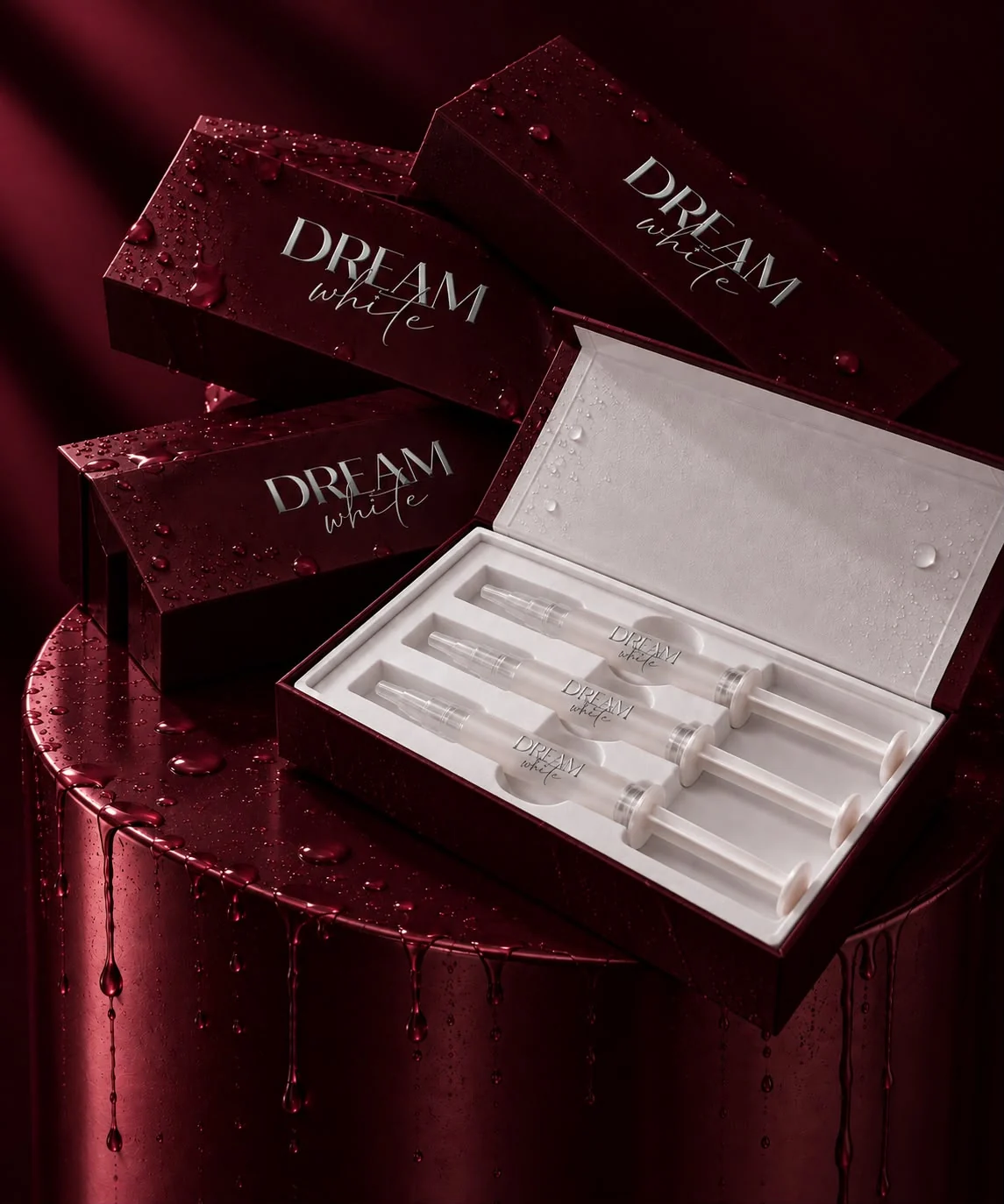 Vier bordeauxfarbene Dreamwhite-Boxen mit Wassertropfen und eine geöffnete Box mit drei Gel-Applikatoren auf einem weinroten Podest