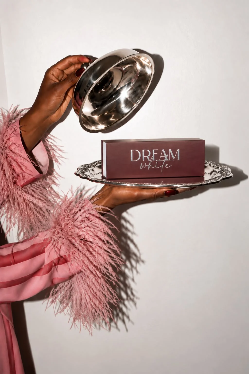 Hand in rosa Federärmel hebt eine silberne Cloche von einer Dreamwhite-Box auf einem Silbertablett